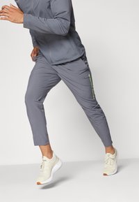 Grå träningskläder som består av en långärmad topp och avsmalnande byxor. Särskild grön branding på sidan. Ljusfärgade sneakers fulländar looken.