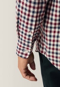 Unterarm und Hand einer Person, die ein rot-weiß-blau kariertes Langarmhemd mit geknöpften Manschetten und dunklen Hosen trägt.