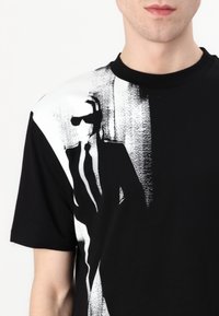 Črna kratkosleeve majica s velikim belim grafičnim posnetkom oblečenega lika. Krpo iz bombaža. Okroglo izrezana vrhnja odprtina. Klasična prilega.