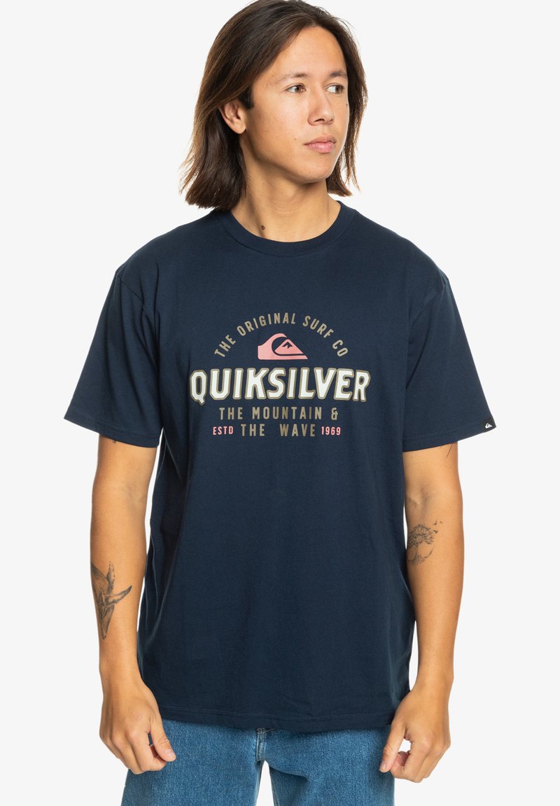 Navy blue cotton t-shirt featuring a Quiksilver logo design in cream and pink, short sleeves, round neckline, and relaxed fit.
