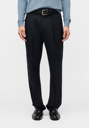 Mand iført sorte bukser med læg, sort bælte, sorte lædersko og en lyseblå striktrøje med lange ærmer.