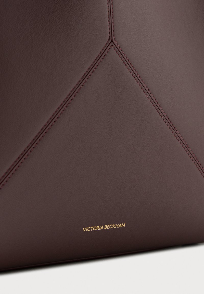 Surface en cuir marron foncé avec des détails de couture triangulaires soignés et un petit texte doré "Victoria Beckham" près du bord inférieur.