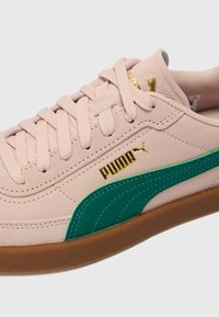 Puma tenisice s gornjim dijelom od blijedo ružičaste kože, zelenim naglascima duž bočne strane i zlatnim detaljem u obliku loga. Smeđa gumena potplat s teksturiranom završnom obradom.