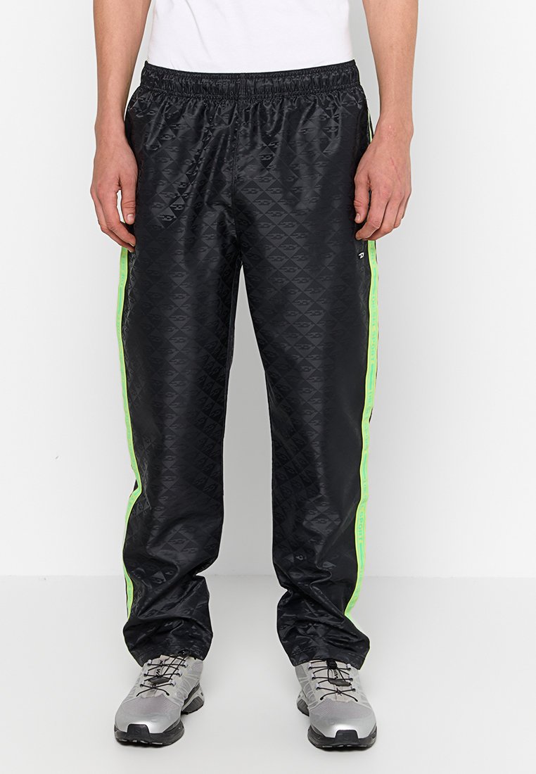 Pantaloni sportivi neri con un motivo a diamante, caratterizzati da strisce laterali fluorescenti verdi e una vita elastica. Abbinati a scarpe da ginnastica grigie.