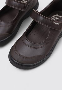 Zapatos de cuero marrón con punta redondeada, correa de Velcro y detalles texturizados. Cuentan con una suela negra flexible y una plantilla perforada para mayor comodidad.