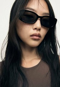 Occhiali da sole neri, rettangolari con montatura spessa, indossati da una modella. Capelli lunghi e scuri e una carnagione liscia con evidenti lentiggini.