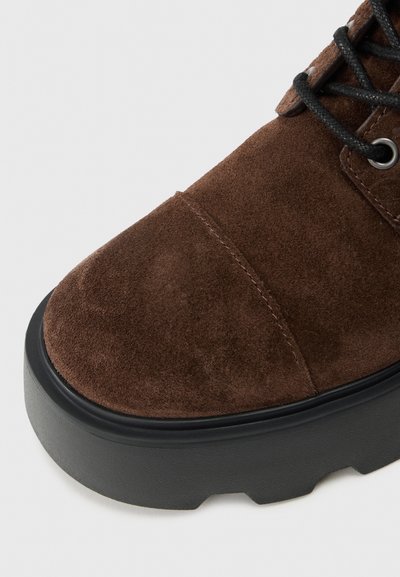 Primo piano di una punta di stivale in suede marrone con suola in gomma nera spessa e lacci neri su sfondo bianco.