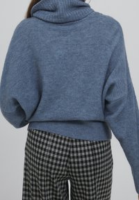 Eine Person in einem lockeren blauen Rollkragenpullover und schwarz-weißen karierten Hosen, von hinten vor einem schlichten grauen Hintergrund gezeigt.