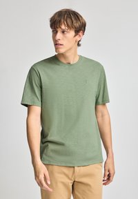 Grön t-shirt med kort ärm gjord av mjukt tyg, med rund halsringning och en liten broderad logotyp på vänster bröst.