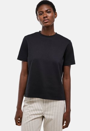 Femme portant un t-shirt noir uni à manches courtes et un pantalon clair avec de fines rayures verticales noires, debout sur un fond blanc.