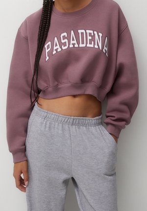 Person trägt mauvefarbenes cropped Sweatshirt mit dem Schriftzug "PASADENA" und hellgraue Jogginghose, mit einer Hand in der Tasche und geflochtenem Haar über der Schulter.