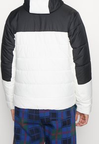 Dunjacka med svart övre del, vit mittenparti och quiltad textur. Har huva och är kombinerad med rutiga byxor.