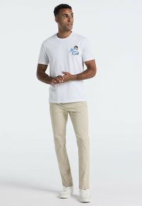 Witte katoenen t-shirt met een multikleurige grafiek op de linkerkant van de borst, gecombineerd met beige broek en witte sneakers. Model staat ontspannen.