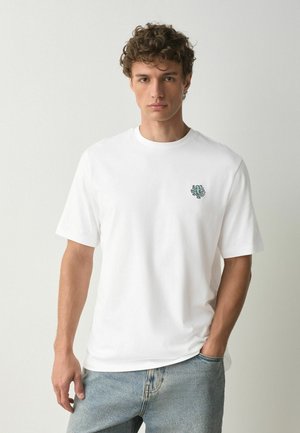Junger Mann mit lockigem Haar, der ein weißes T-Shirt mit einem kleinen abstrakten Logo auf der linken Brustseite und hellblaue Jeans trägt.
