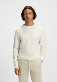 Sudadera de cuello redondo texturizada en beige claro con un sutil patrón acanalado, adorned con un pequeño logo en el pecho y puños ajustados.