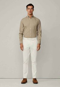 Άνδρας στέκεται με πρόσωπο προς τα εμπρός φορώντας μπεζ πουκάμισο με κουμπιά, λευκά παντελόνια και καφέ δερμάτινα παπούτσια σε απλό ανοιχτό γκρι φόντο.