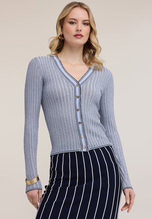 Blonde Frau, die eine hellblaue, gerippte, geknöpfte Strickjacke und einen marineblauen Rock mit vertikalen weißen Streifen trägt, posiert vor einem beigen Hintergrund.