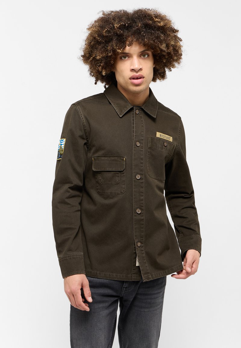 Camicia verde oliva con bottoni realizzata in un tessuto resistente, caratterizzata da una tasca sul petto, una patch con logo dorato e un accento a fantasia sulle maniche.
