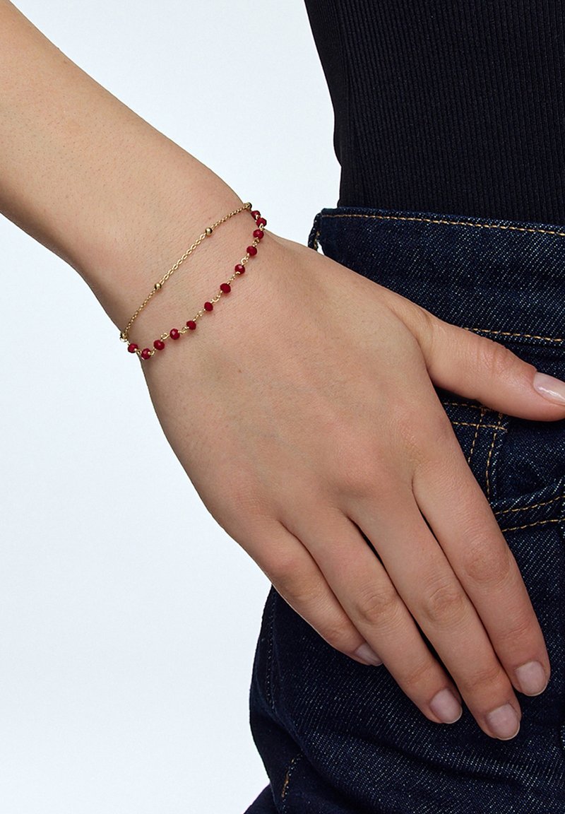 Złota bransoletka z czerwonymi koralikami, o delikatnym wzornictwie. Gładka tekstura i okrągłe akcenty nadają elegancki wygląd.