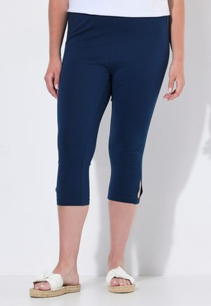 Vrouw draagt marineblauwe cropped leggings en witte instapsandalen met een gevlochten zool, staand tegen een effen lichte achtergrond.