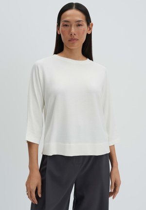 Donna con lunghi capelli scuri che indossa una maglia bianca a maniche tre quarti con texture e pantaloni neri larghi, in piedi contro uno sfondo chiaro.