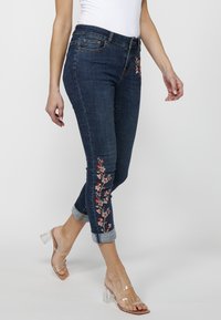 Jeans de mezclilla azul oscuro con diseños florales bordados en rojo y rosa a lo largo de los lados, con puños enrollados y una forma ajustada.