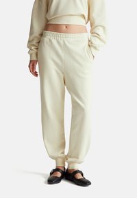 Jasno beżowe dresy z elastycznym paskiem i dopasowanymi mankietami. Miękka faktura i luźny krój, zestawione z czarnymi płaskimi butami z akcentami pasków.