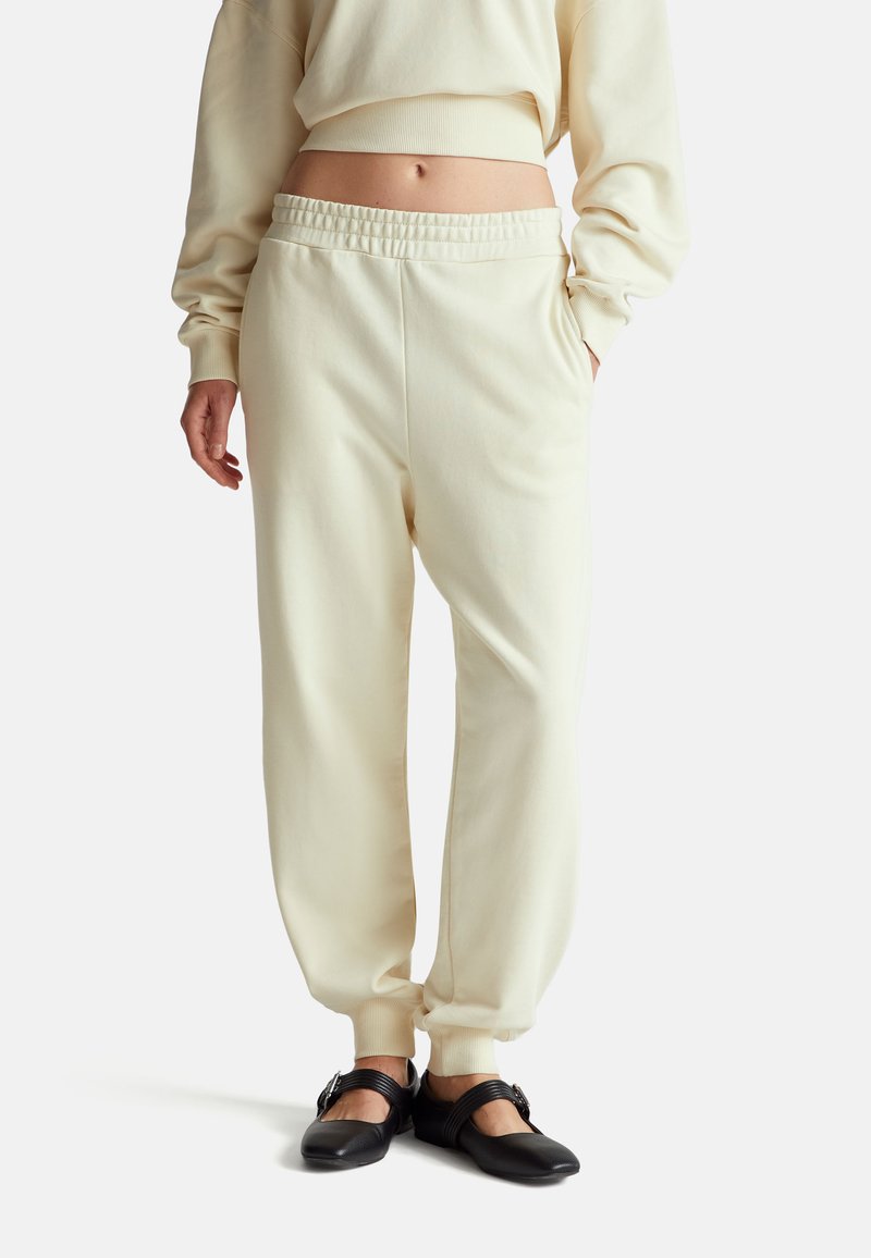 Jasno beżowe dresy z elastycznym paskiem i dopasowanymi mankietami. Miękka faktura i luźny krój, zestawione z czarnymi płaskimi butami z akcentami pasków.