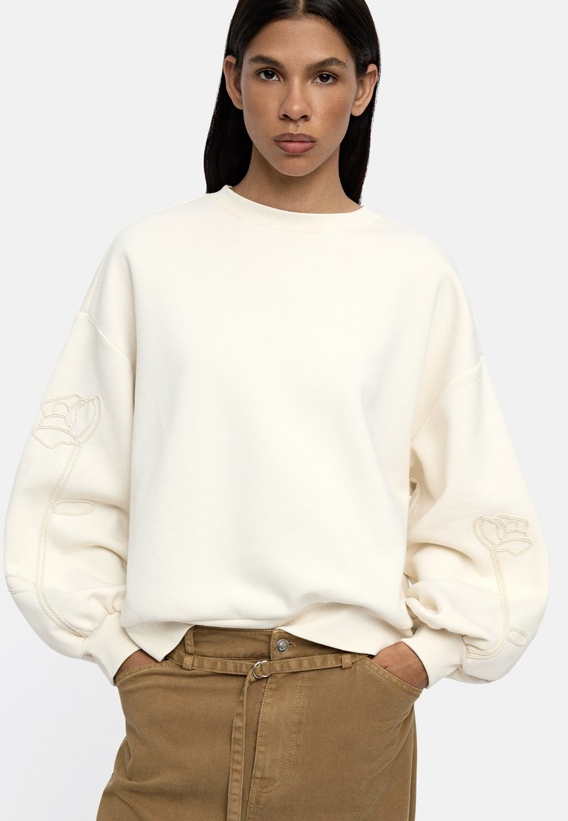 Kremowy sweter z oversize'owymi rękawami, z haftowanym wzorem kwiatowym na lewej ręce. Miękka faktura, luźny krój.