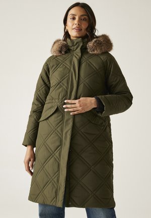 Kobieta w oliwkowozielonym pikowanym płaszczu z kapturem obszytym futerkiem, stojąca z jedną ręką na przedniej części płaszcza, na jednolitym tle.