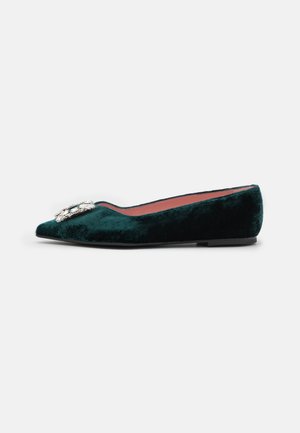 Zapato plano de terciopelo verde con punta afilada, que presenta un broche decorativo de cristal en la parte delantera y una suela negra plana.