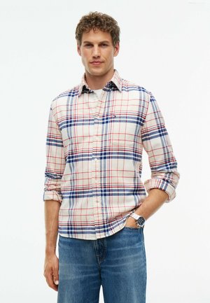 Mand med krøllet hår iført en hvid, blå og rød ternet skjorte med lange ærmer, blå jeans og et armbåndsur, stående med den ene hånd i lommen.