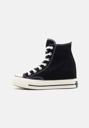 Zwarte hoge canvas sneaker met witte veters, witte rubberen zool en zwarte streepdetail tegen een effen lichte achtergrond.
