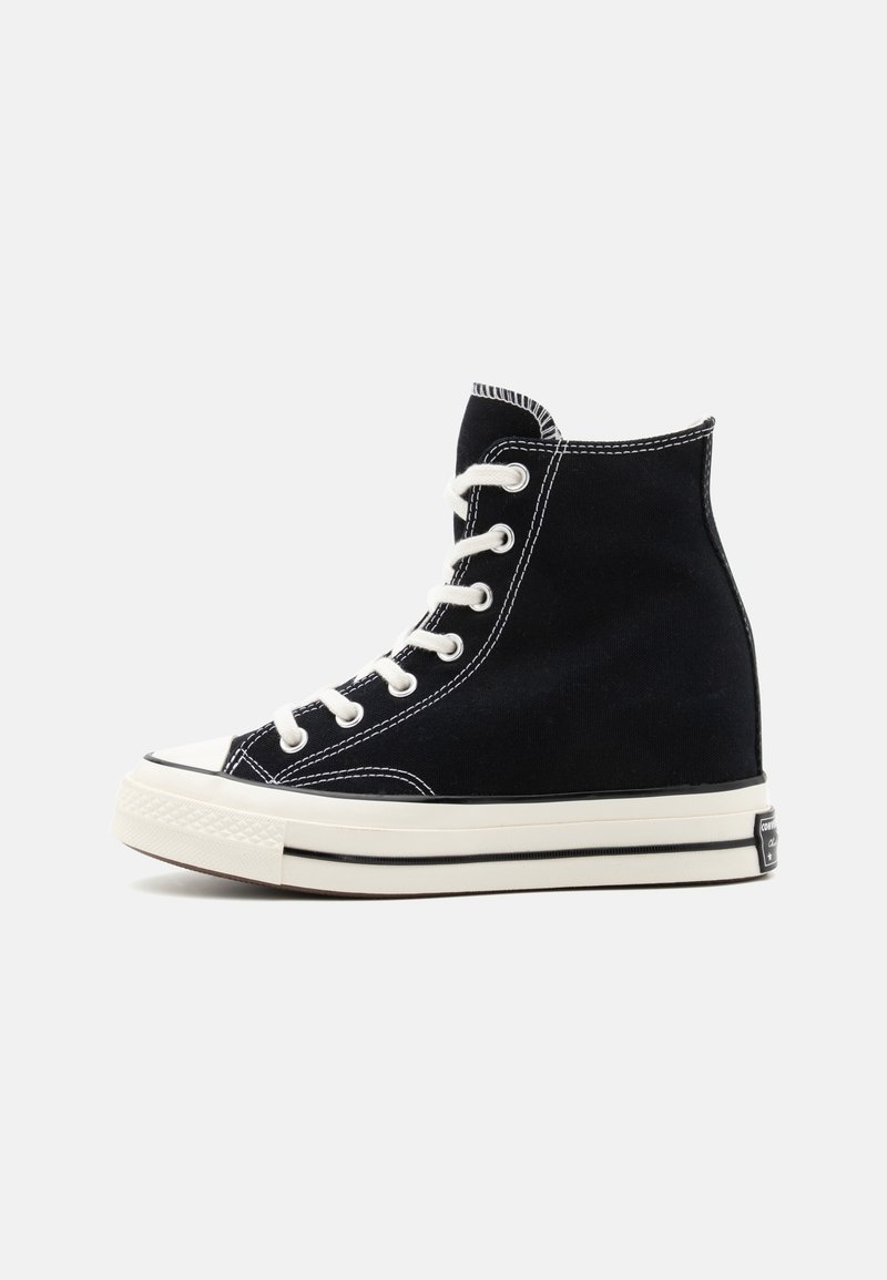 Baskets montantes en toile noire avec lacets blancs, semelle en caoutchouc blanche et détail d'une bande noire sur un fond clair uni.
