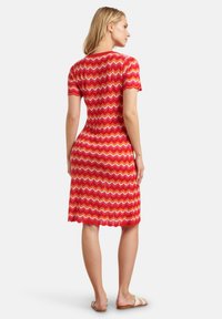 Rood gekleurde jurk met zigzagpatroon, korte mouwen, knielange snit en ribbelstructuur. Model draagt lichte sandalen en staat met de rug naar de camera.