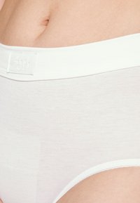 Hvit bomullsbrief med en myk og glatt tekstur. Har en bred midjebånd med brodert logo. Enkelt, klassisk design.