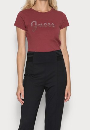 Femme portant un t-shirt ajusté bordeaux avec un logo "Guess" en strass et un pantalon noir taille haute avec des panneaux de ceinture texturés.