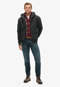 Sudadera con cremallera de color gris oscuro, forrada de felpa, llevada sobre una camisa de cuadro roja, jeans azules y botas de cordones marrones.