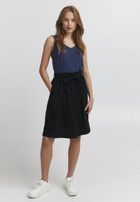 Navy mouwloze top, gecombineerd met een zwarte rok tot de knie met een elastische tailleband en strikdetail, afgemaakt met witte sneakers.