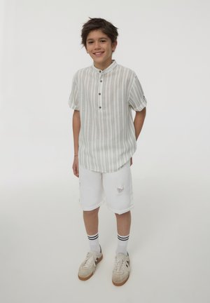 Camicia a maniche corte con lievi righe e colletto abbottonato, abbinata a pantaloni corti bianchi al ginocchio con bordi sfilacciati. Footwear chiaro e calze a righe.