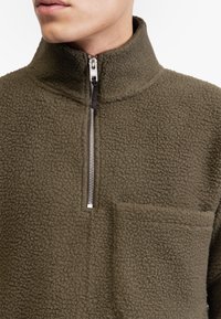 Olivgrüner Fleece-Pullover mit hohem Kragen, silbernem Reißverschluss und einer Fronttasche. Strukturierter, weicher Stoff mit einem gemütlichen Aussehen.