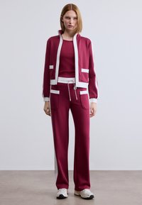 Μπορντό αθλητικό σετ φόρμας με μπουφάν με φερμουάρ και λευκές λεπτομέρειες, καθώς και παντελόνι με ρίγες στα πλάγια, κατασκευασμένο από απαλό, ελαστικό ύφασμα.