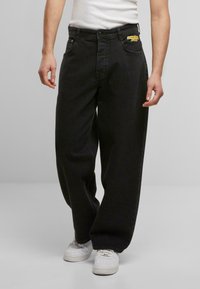 Schwarze Denim-Hosen mit lockerem Schnitt, versehen mit einem gelben gestickten Logo am Bund und Fronttaschen. Getragen mit weißen Sneakers.