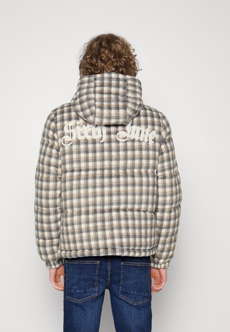 Person med lockigt hår som bär en grå och beige rutmönstrad puffjacka med huva och texten "Scaly Lore" på baksidan, i kombination med blå jeans.