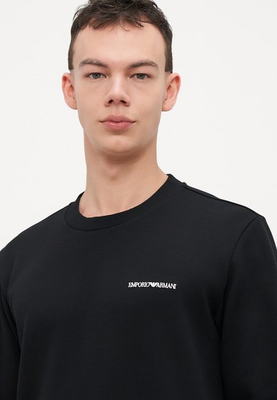 Sweat-shirt noir à manches longues avec un col rond, présentant un petit logo brodé blanc "EMPORIO ARMANI" sur la poitrine.