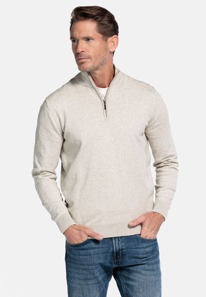 Hellbeiger Pullover mit Reißverschluss-Kragen, gefertigt aus weichem Material, mit langen Ärmeln und gerippten Bündchen, kombiniert mit blauer Jeans.