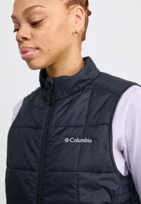 Zwarte geïsoleerde vest met een hoge kraag, gestikte secties en een rits aan de voorkant. Bevat het Columbia-logo in het wit op de linkerkant van de borst.