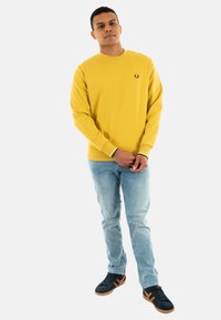 Sudadera amarilla de manga larga y cuello redondo, con un pequeño logo negro en el pecho. Se combina con jeans de color azul claro y zapatillas negras.