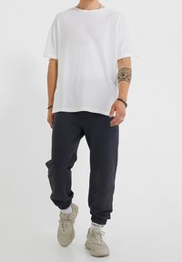 Muž v volném bílém tričku, černých teplákových kalhotách, bílých ponožkách, béžových teniskách, s kmenovým tetováním na pravém předloktí a náramky na obou zápěstích.