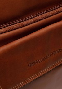 Borsa in pelle marrone con una texture liscia e cuciture finemente lavorate; chiusura con zip; logo impresso e accenti sottili lungo i bordi.